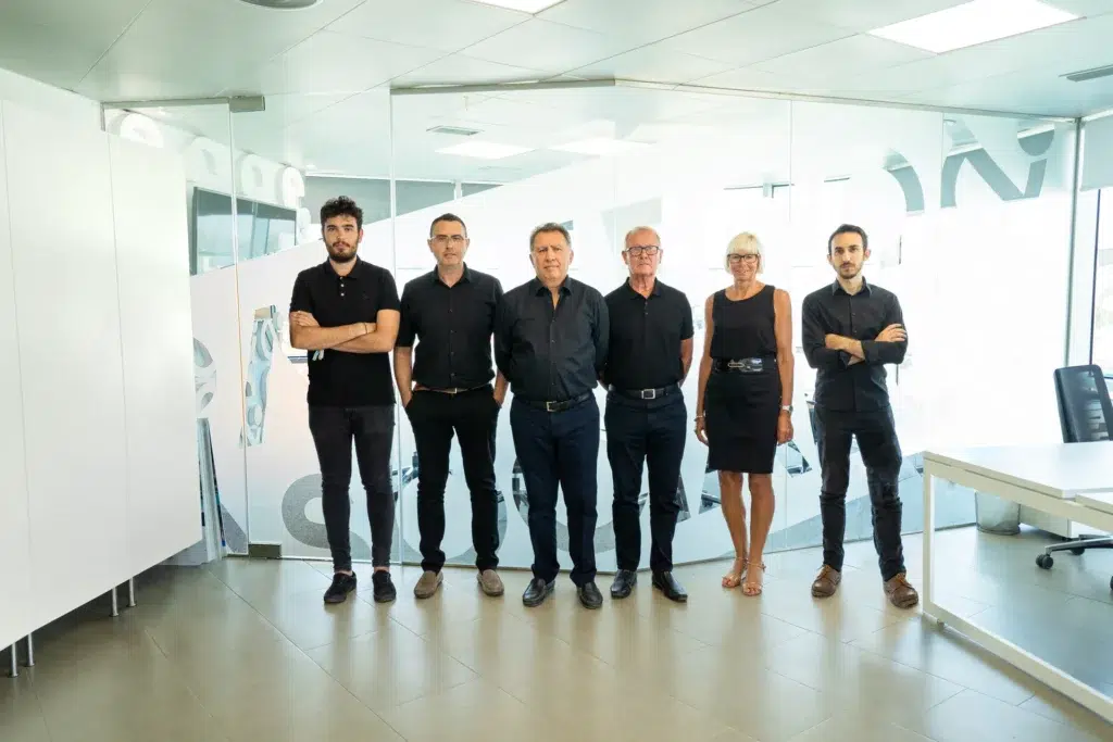 The Pacheco Architects team standing in a line inside their office, posing in front of a glass wall with the firm's branding.