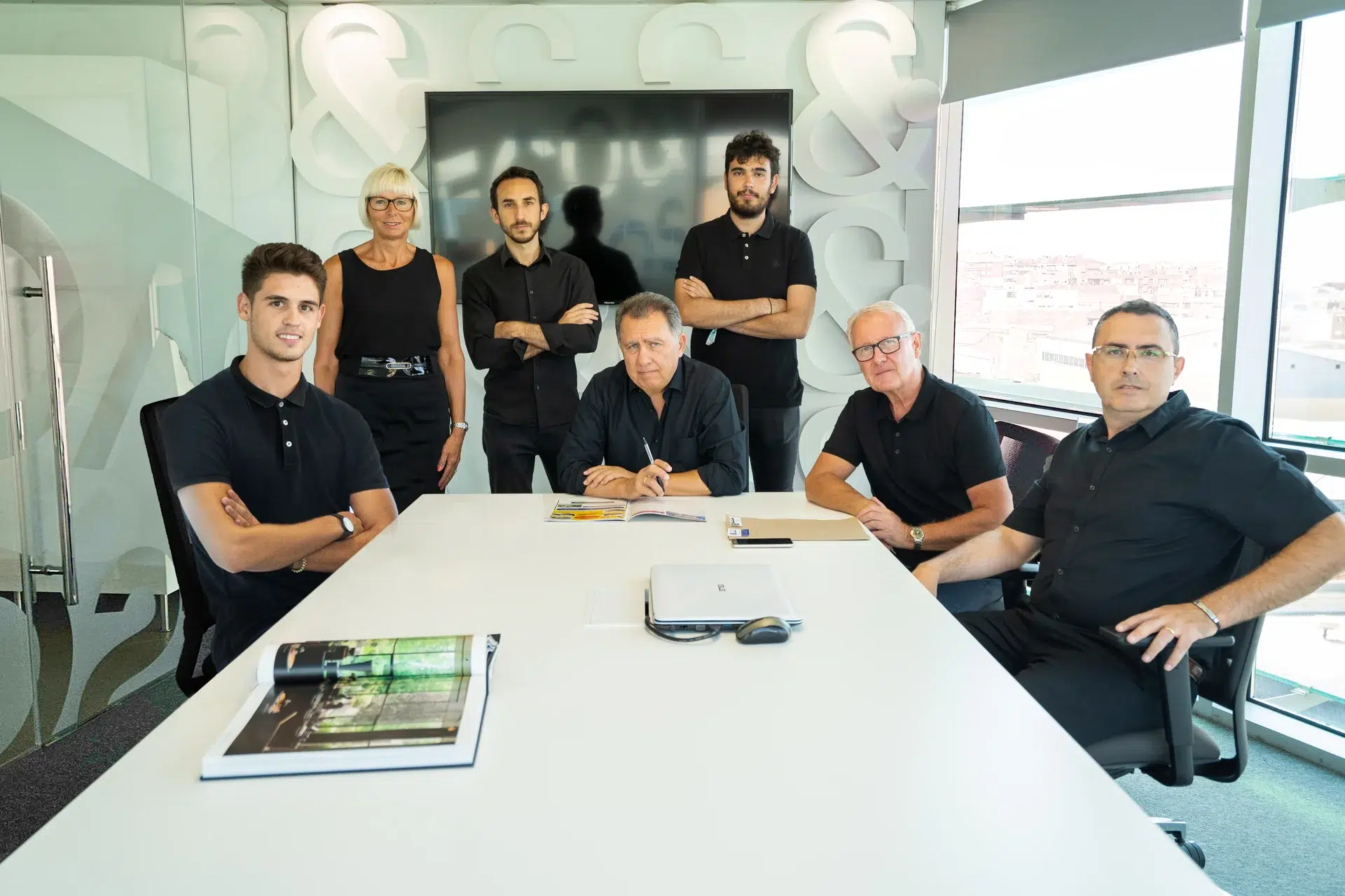 The Pacheco Architects team gathered around a meeting table, with documents and a laptop, in a modern office setting.