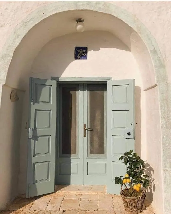 Elegant pastel green wooden door framed by a whitewashed arched wall and a basket of lemons, typical of Mediterranean vernacular housing.